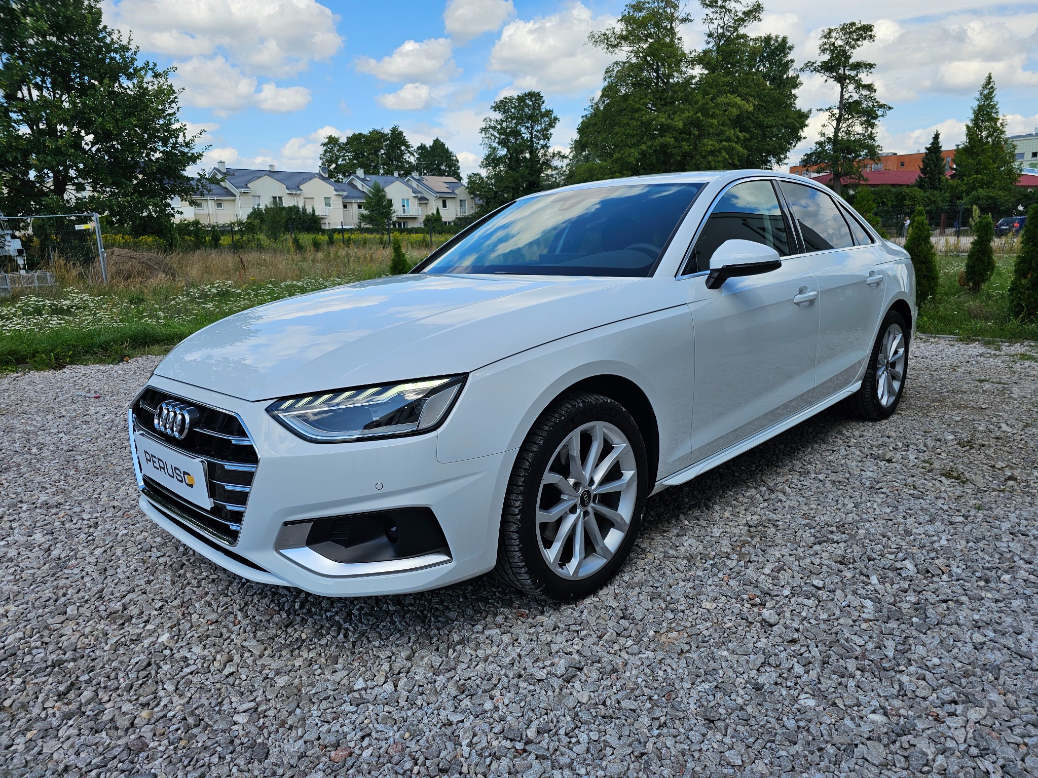 Biała limuzyna Audi A4 na żwirowym podłożu, widok z przodu, w tle domy jednorodzinne i drzewa pod błękitnym niebem z chmurami.