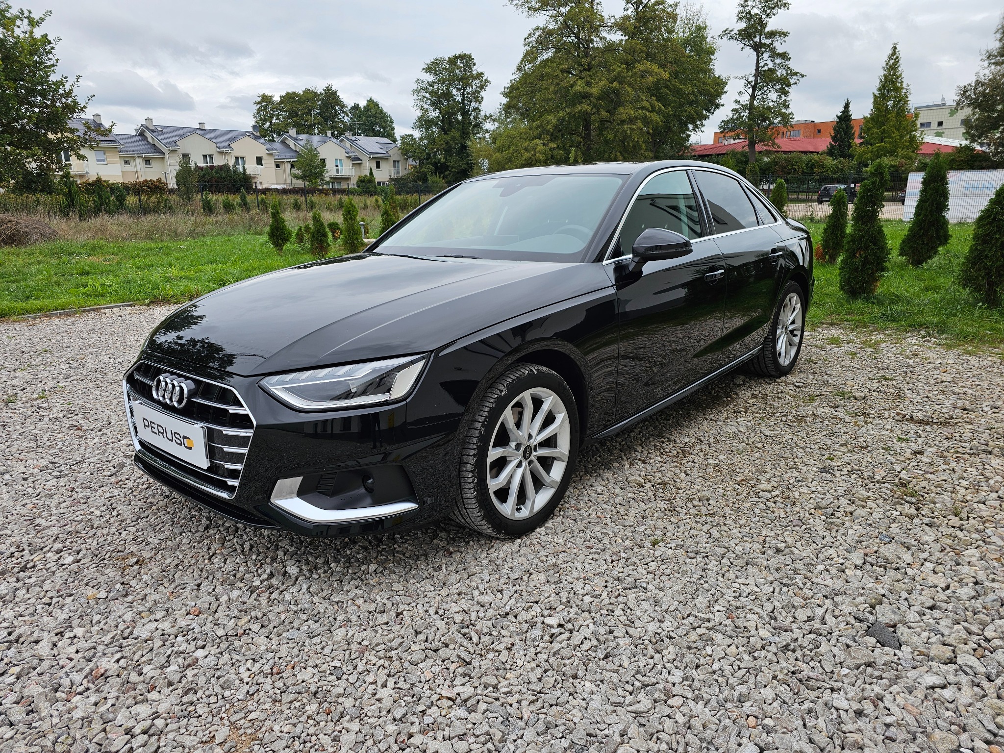 Czarny sedan Audi A4 z widocznym logo firmy Peruso na tablicy rejestracyjnej, zaparkowany na żwirowym podłożu, z budynkami mieszkalnymi w tle.