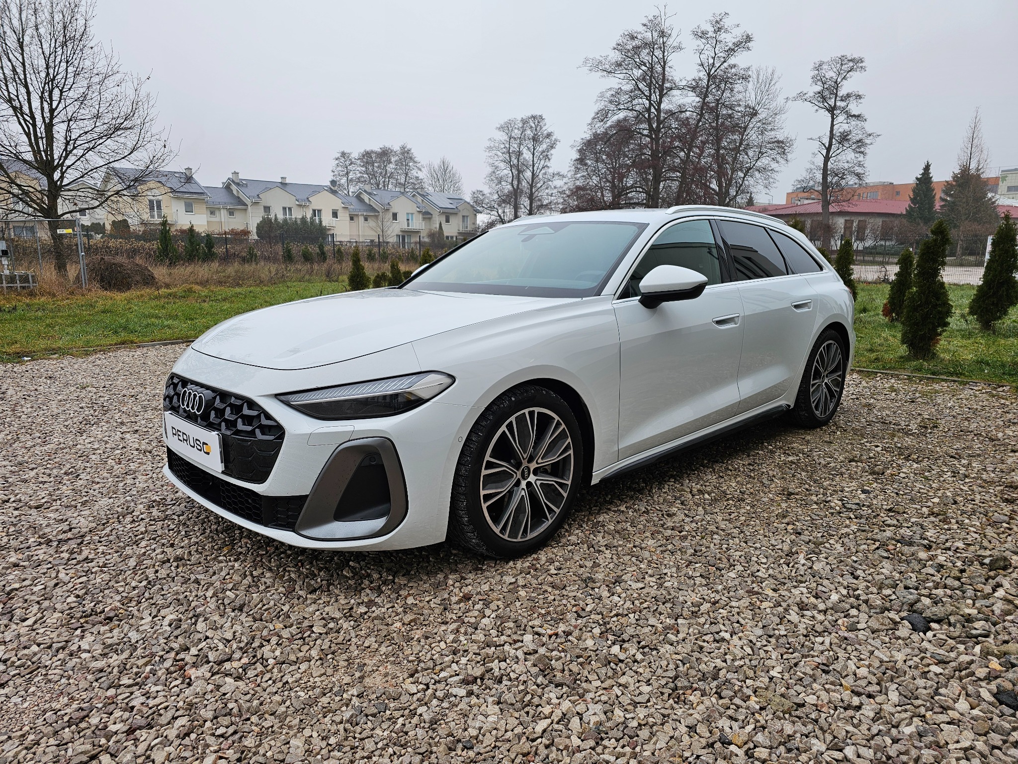 Białe Audi A6 Avant na żwirowym podłożu w Warszawie, z widocznym logo firmy na tablicy rejestracyjnej i budynkami mieszkalnymi w tle.