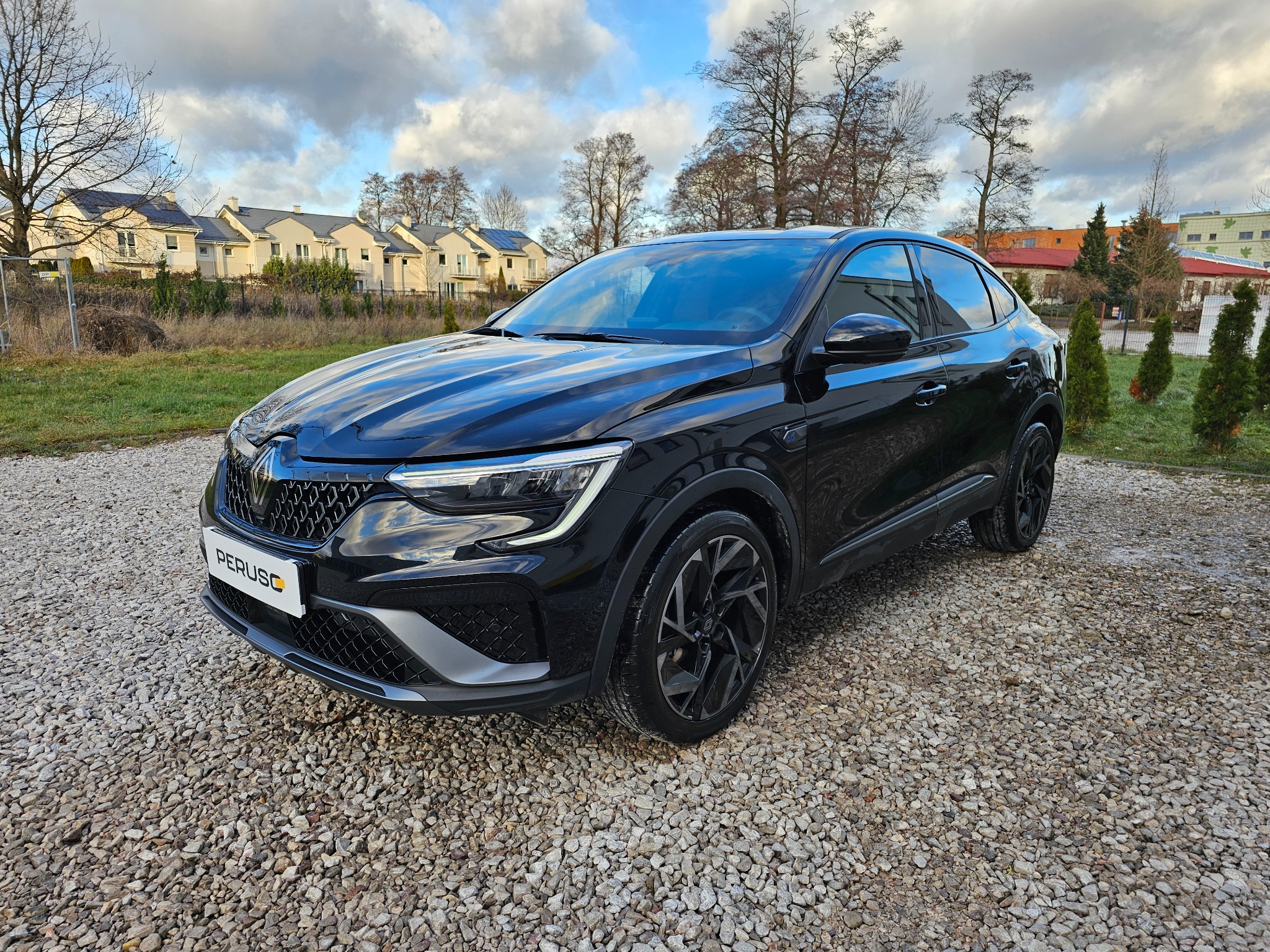 Czarny samochód typu SUV marki Renault, zaparkowany na żwirowym podłożu, z widocznym logo firmy na tablicy rejestracyjnej, na tle osiedla domów jednorodzinnych i pochmurnego nieba.