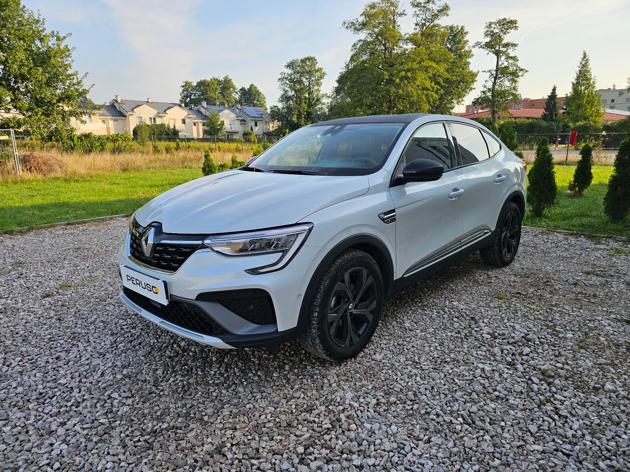 Biały samochód Renault Arkana z czarnymi felgami, zaparkowany na żwirowym podjeździe przed domem w słoneczny dzień.