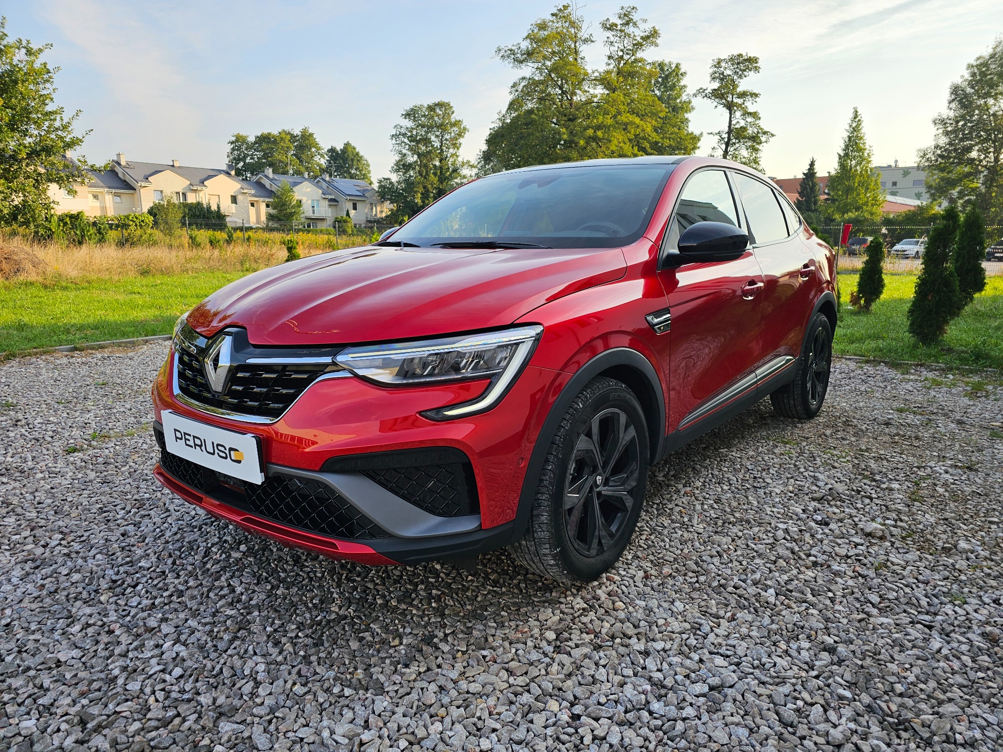 Czerwony samochód Renault Arkana z czarnymi felgami stoi na żwirowym podłożu, w tle domy jednorodzinne i drzewa, oświetlony popołudniowym słońcem.