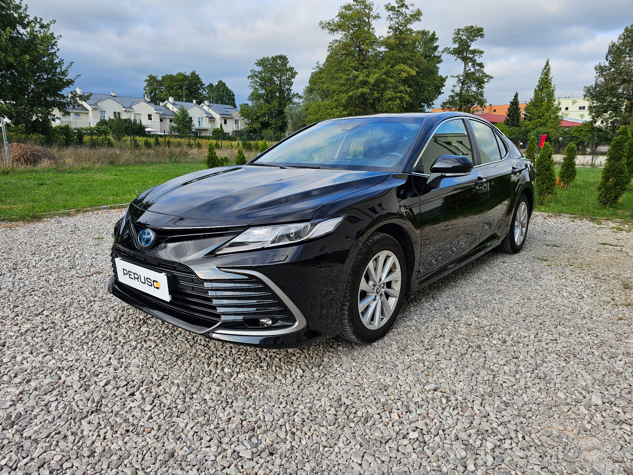 Czarny, lśniący samochód hybrydowy marki Toyota Camry, zaparkowany na żwirowym podłożu, z widocznym logo firmy PERUSC na przedniej tablicy rejestracyjnej, na tle budynków mieszkalnych i zieleni.