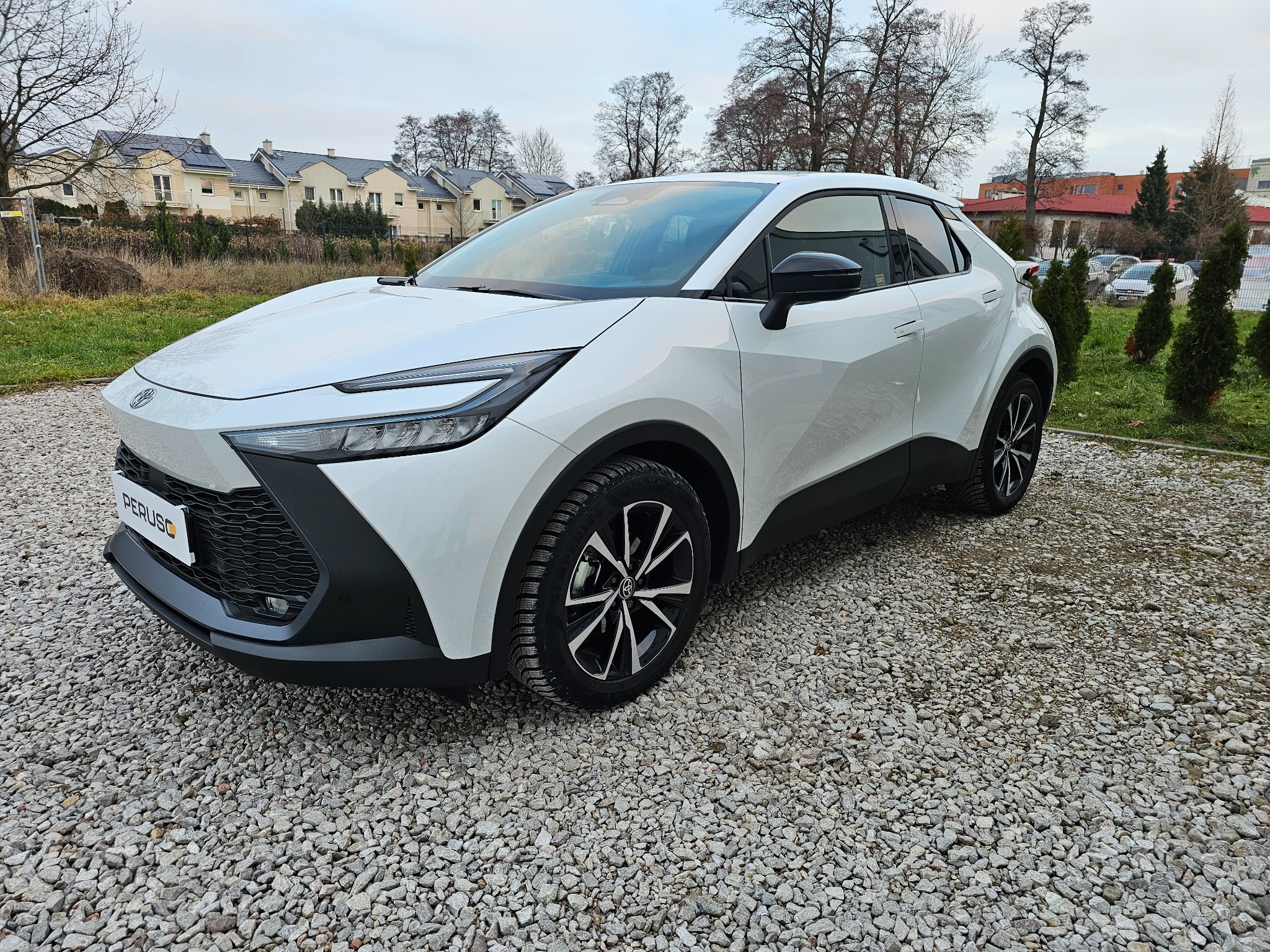 Biały samochód typu crossover z czarnymi elementami wykończenia, zaparkowany na żwirowym podłożu przed budynkami mieszkalnymi w pochmurny dzień.