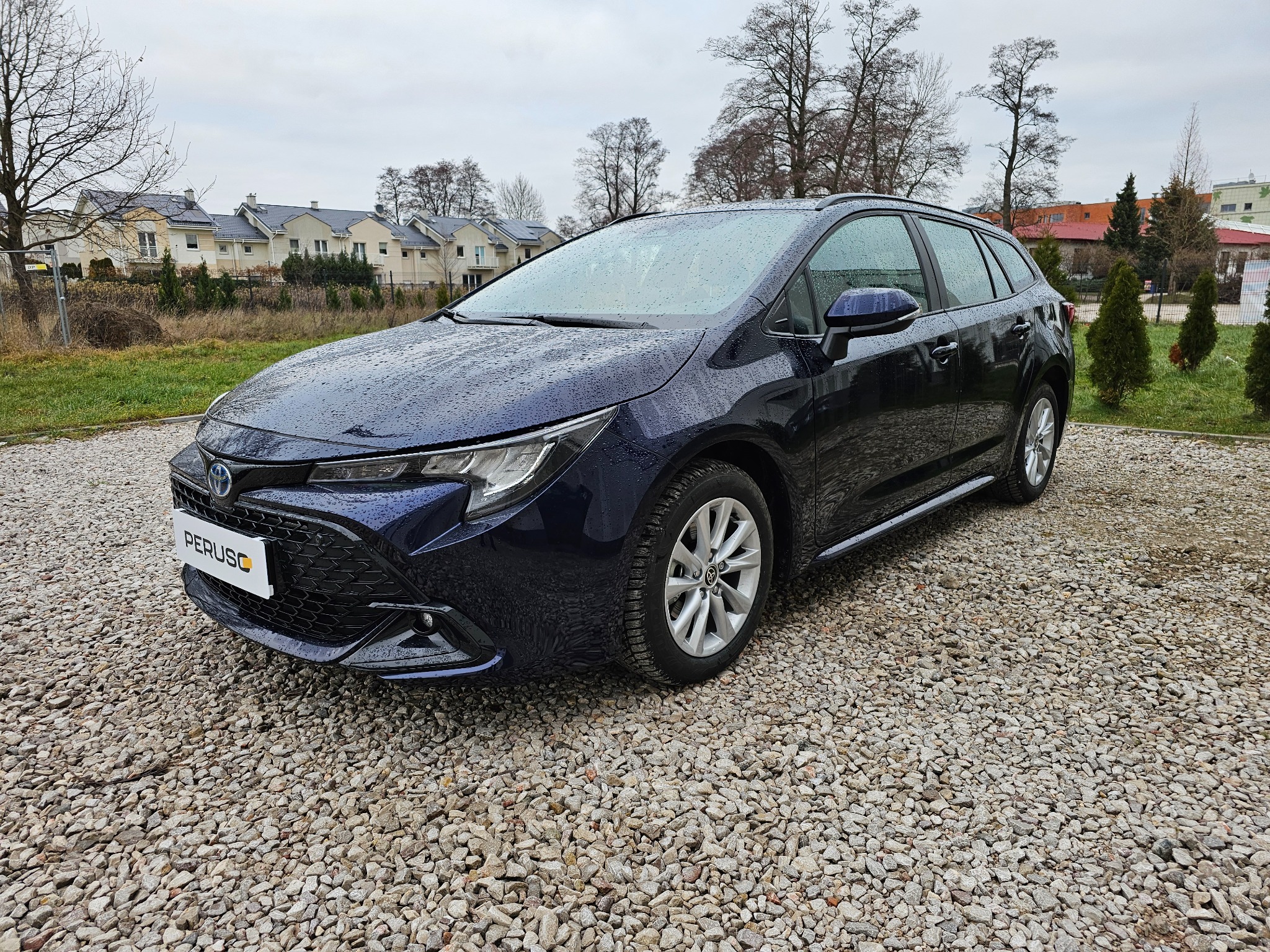 Granatowa Toyota Corolla kombi na żwirowym podłożu, widoczne krople deszczu na karoserii, w tle budynki mieszkalne i drzewa.