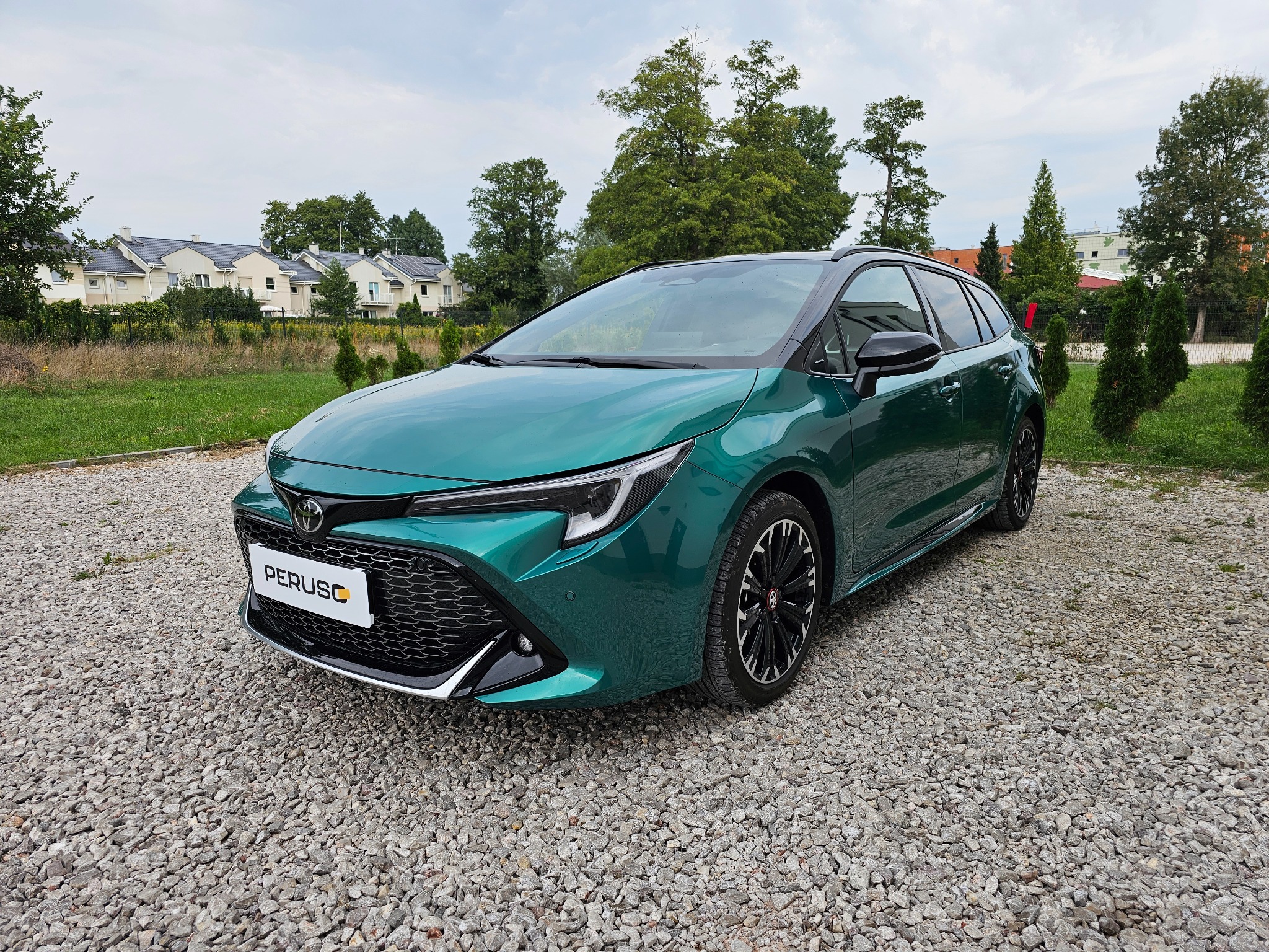 Szmaragdowy samochód kombi z czarnymi felgami parkuje na żwirowym podłożu przed budynkami mieszkalnymi i drzewami w pochmurny dzień.