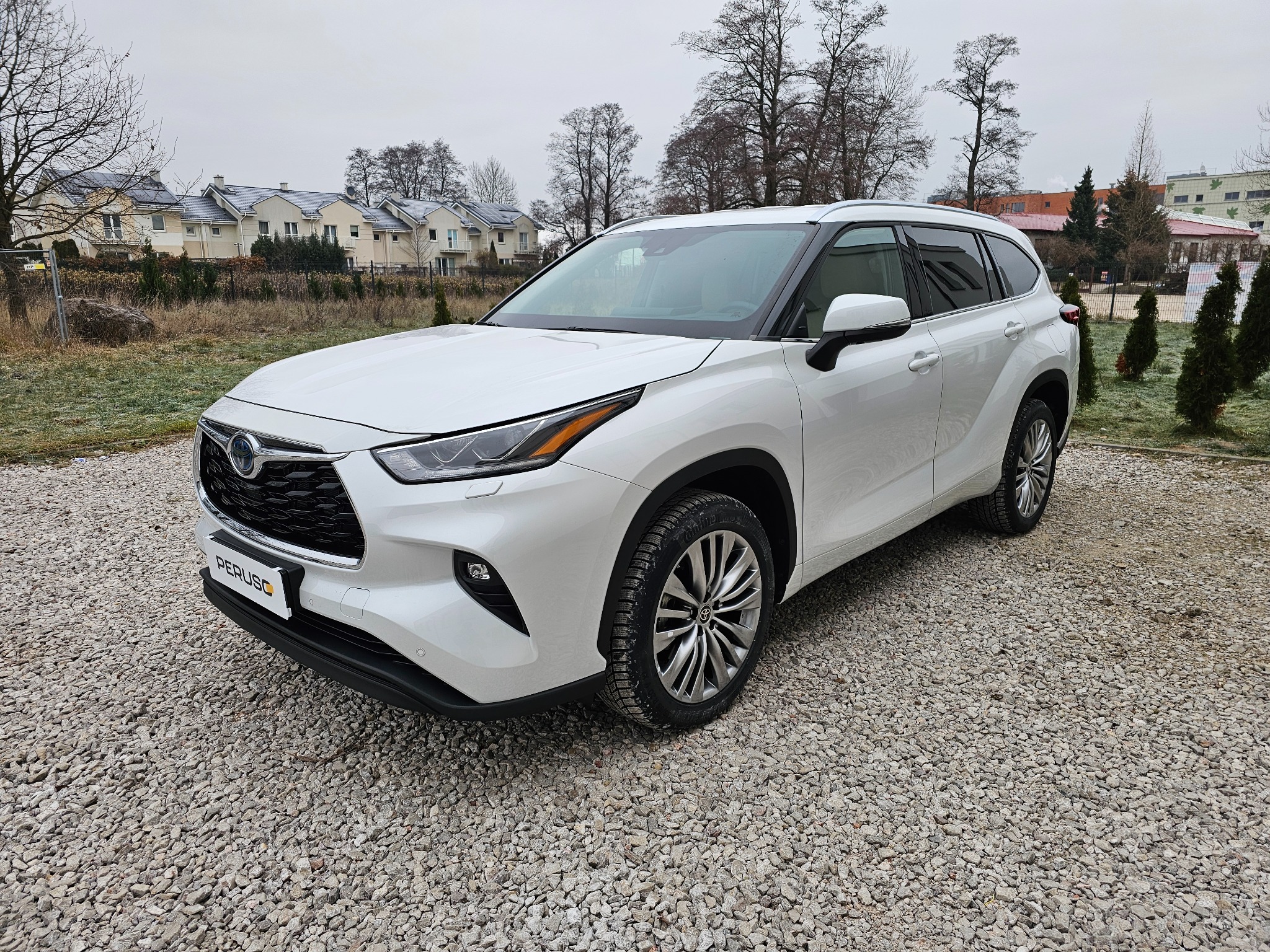 Biały SUV Toyota Highlander zaparkowany na żwirowym podłożu, widok z przodu, w tle osiedle domów jednorodzinnych.