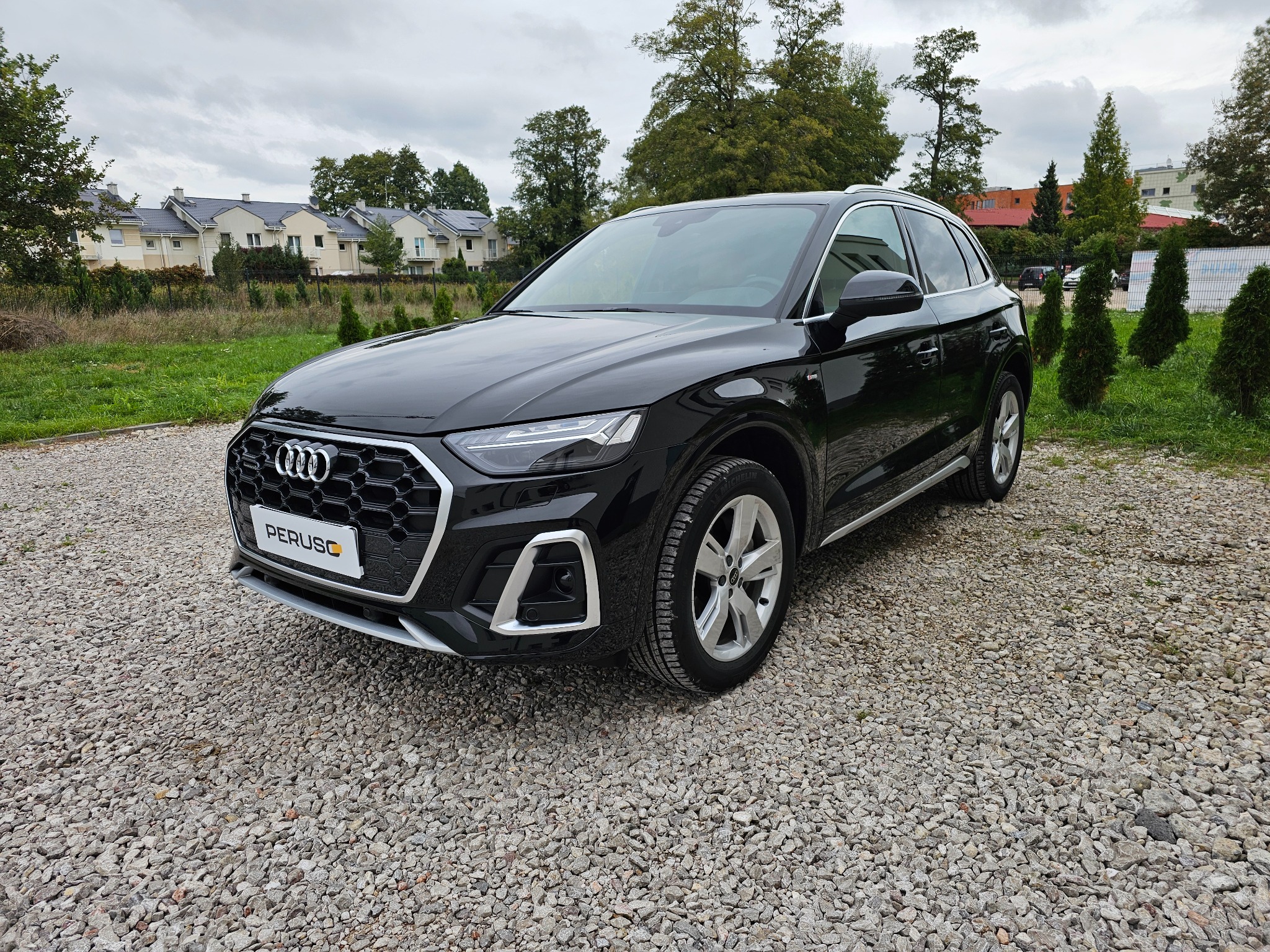 Czarny samochód Audi Q5 na żwirowym podłożu, z widocznym logo dealera 'PERUSC' na przedniej tablicy rejestracyjnej, w tle zabudowa jednorodzinna i zieleń.