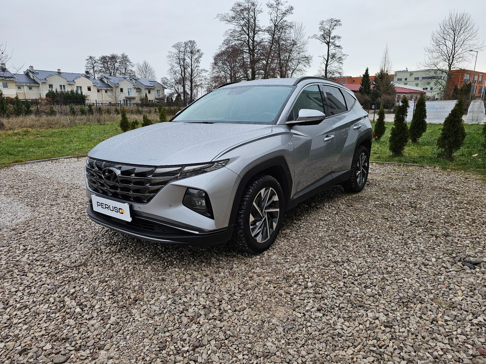Srebrny Hyundai Tucson stoi na żwirowym parkingu, widoczne krople deszczu na karoserii, w tle budynki mieszkalne i drzewa.
