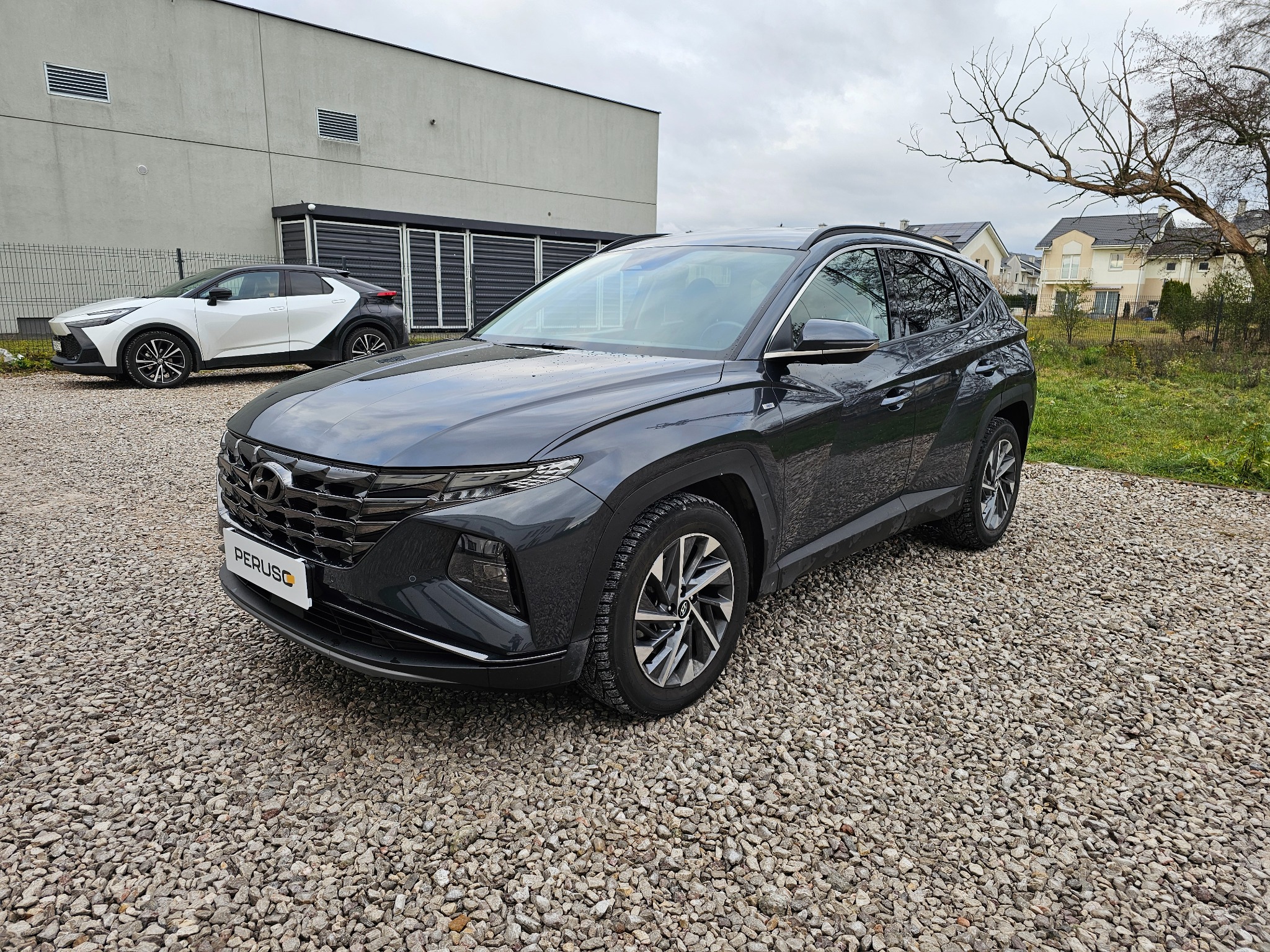 Ciemnoszary samochód Hyundai Tucson na żwirowym parkingu przed nowoczesnym budynkiem, z widoczną tablicą rejestracyjną 'PERUSC' i białym samochodem w tle.