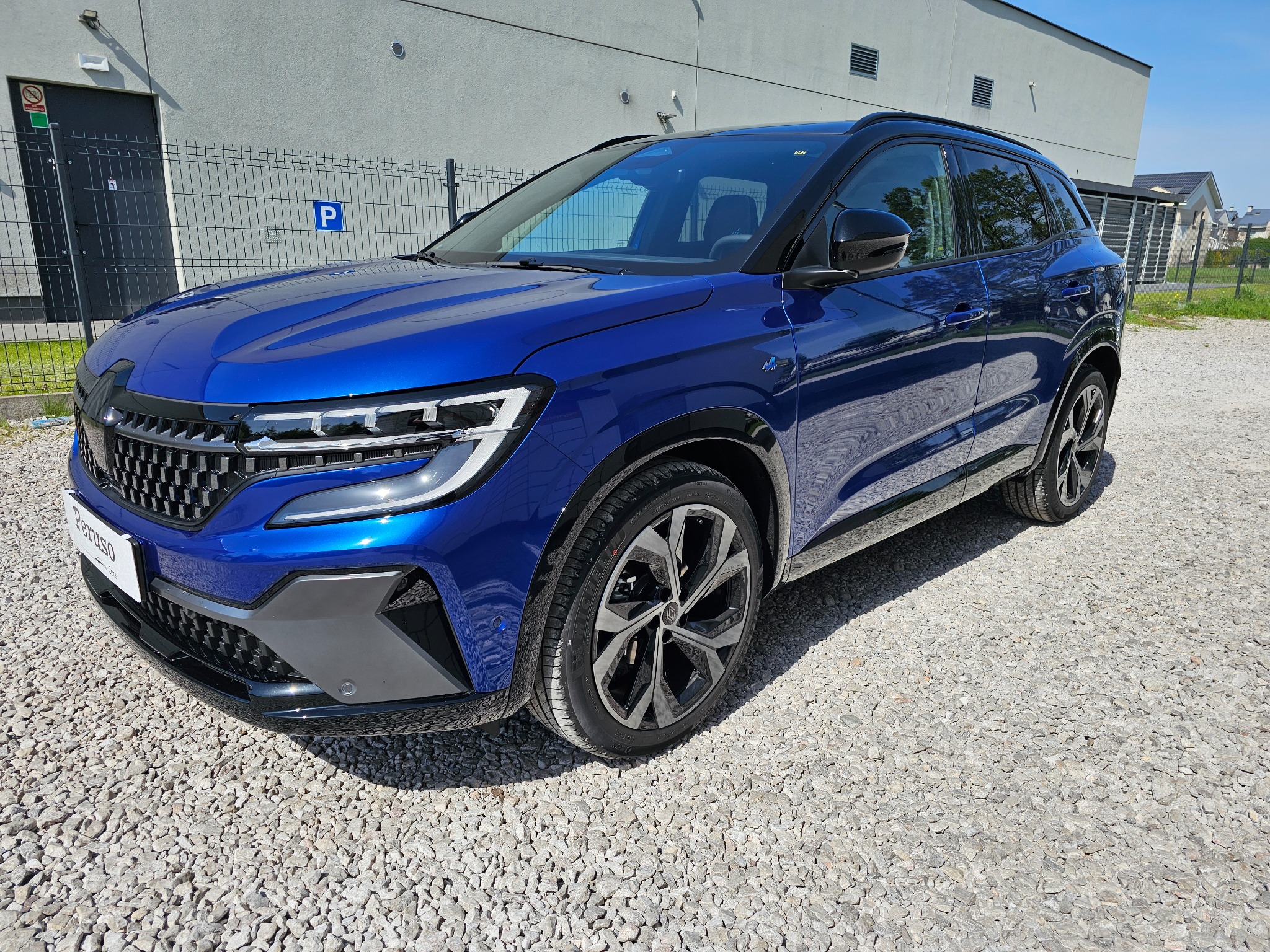 Niebieski samochód Renault Austral na żwirowym parkingu przed szarym budynkiem z oznaczeniem parkingu.