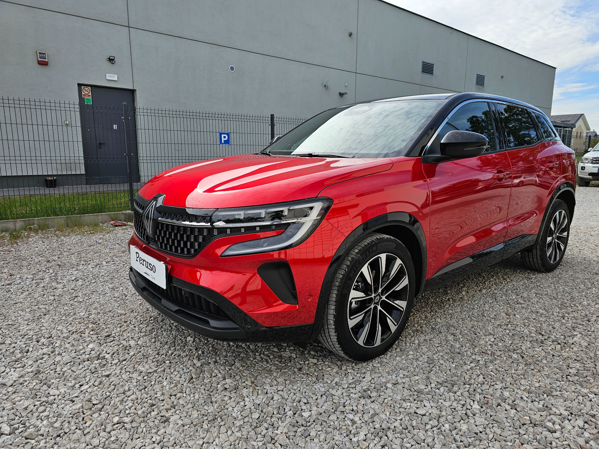 Czerwony samochód typu SUV marki Renault, zaparkowany na żwirowym parkingu przed szarym budynkiem z niebieskim znakiem parkingowym.