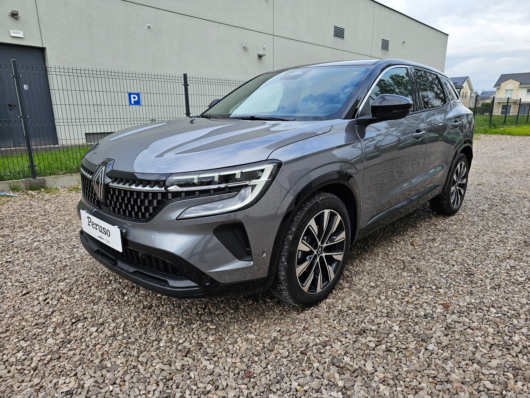 Szary samochód Renault na żwirowym parkingu przed budynkiem z szarą elewacją i znakiem parkingu w tle.