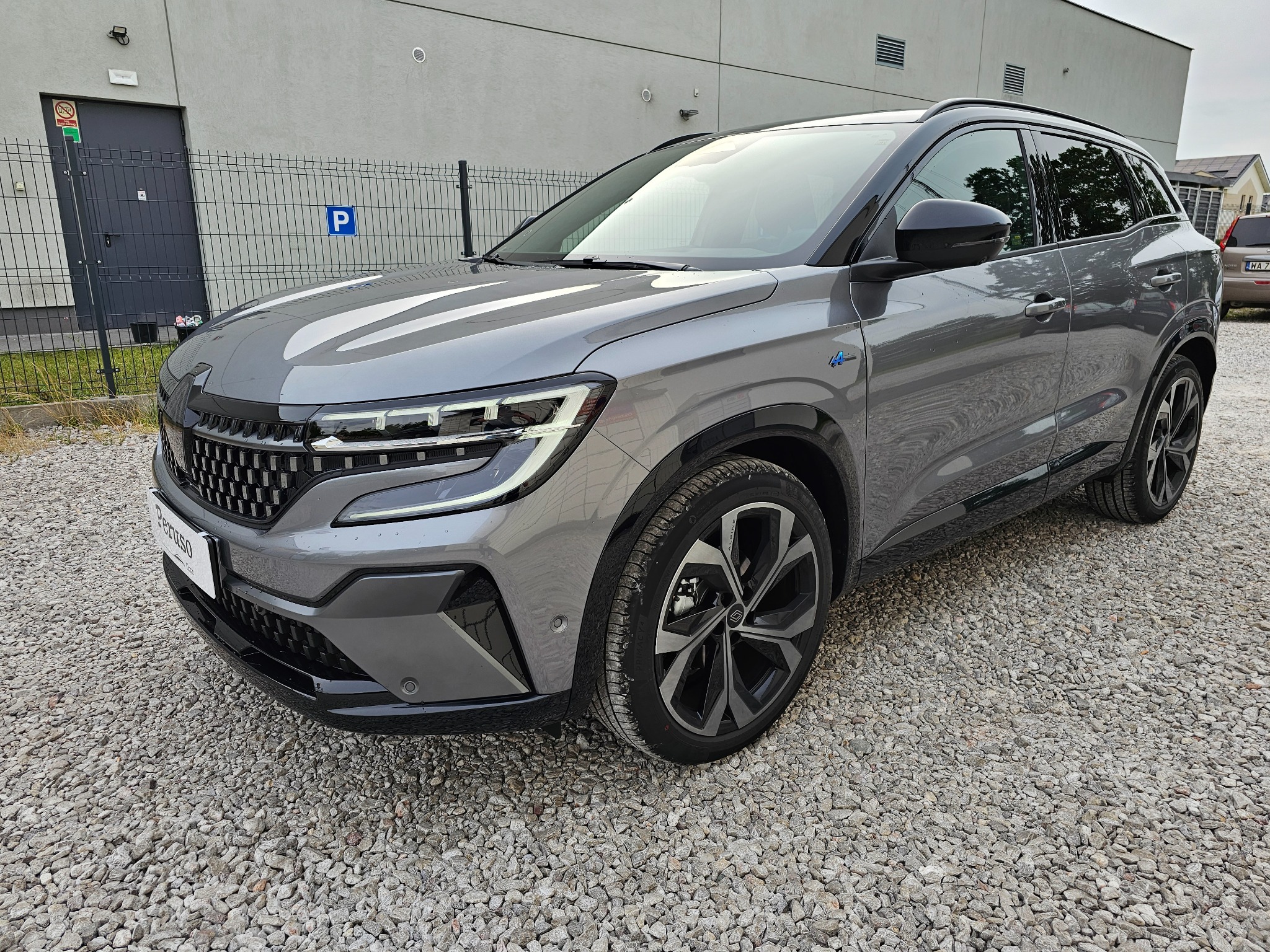 Szary SUV Renault Austral E-Tech w wersji hybrydowej, zaparkowany na żwirowym parkingu przed budynkiem z szarą elewacją i znakiem parkingu.