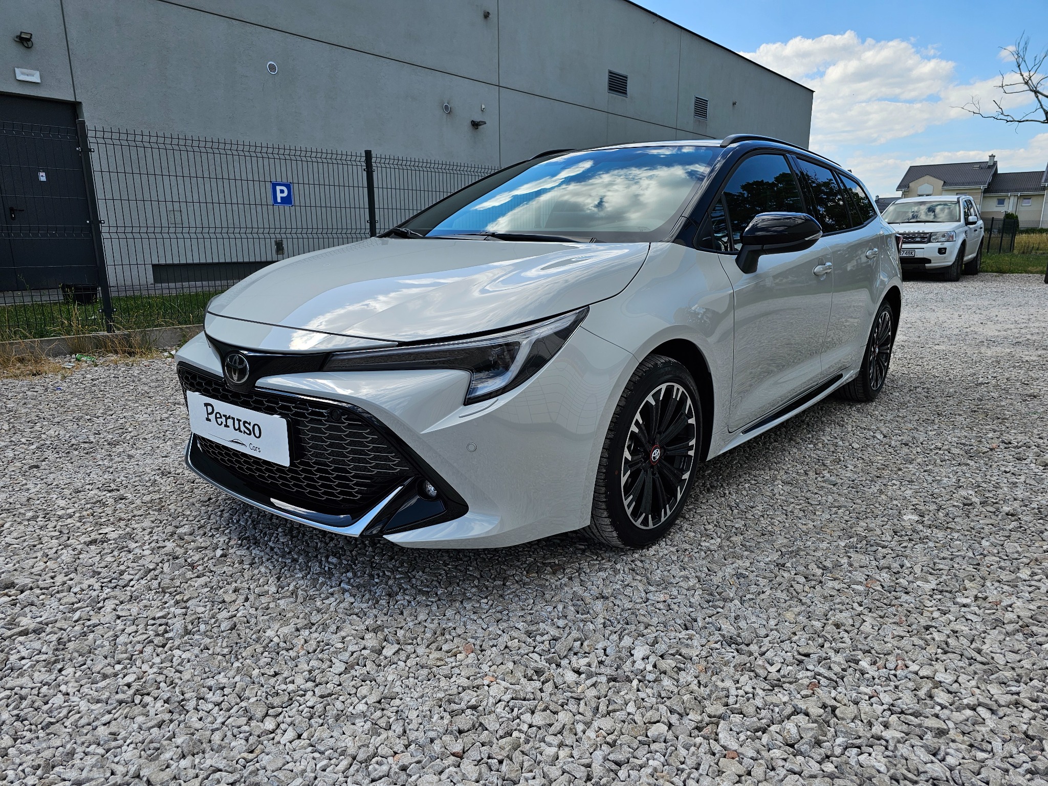 Jasnoszary samochód kombi na żwirowym parkingu, z widocznym logo 'Peruso Cars' na przedniej tablicy rejestracyjnej, zaparkowany przed szarym budynkiem z ogrodzeniem i znakiem 'P'.