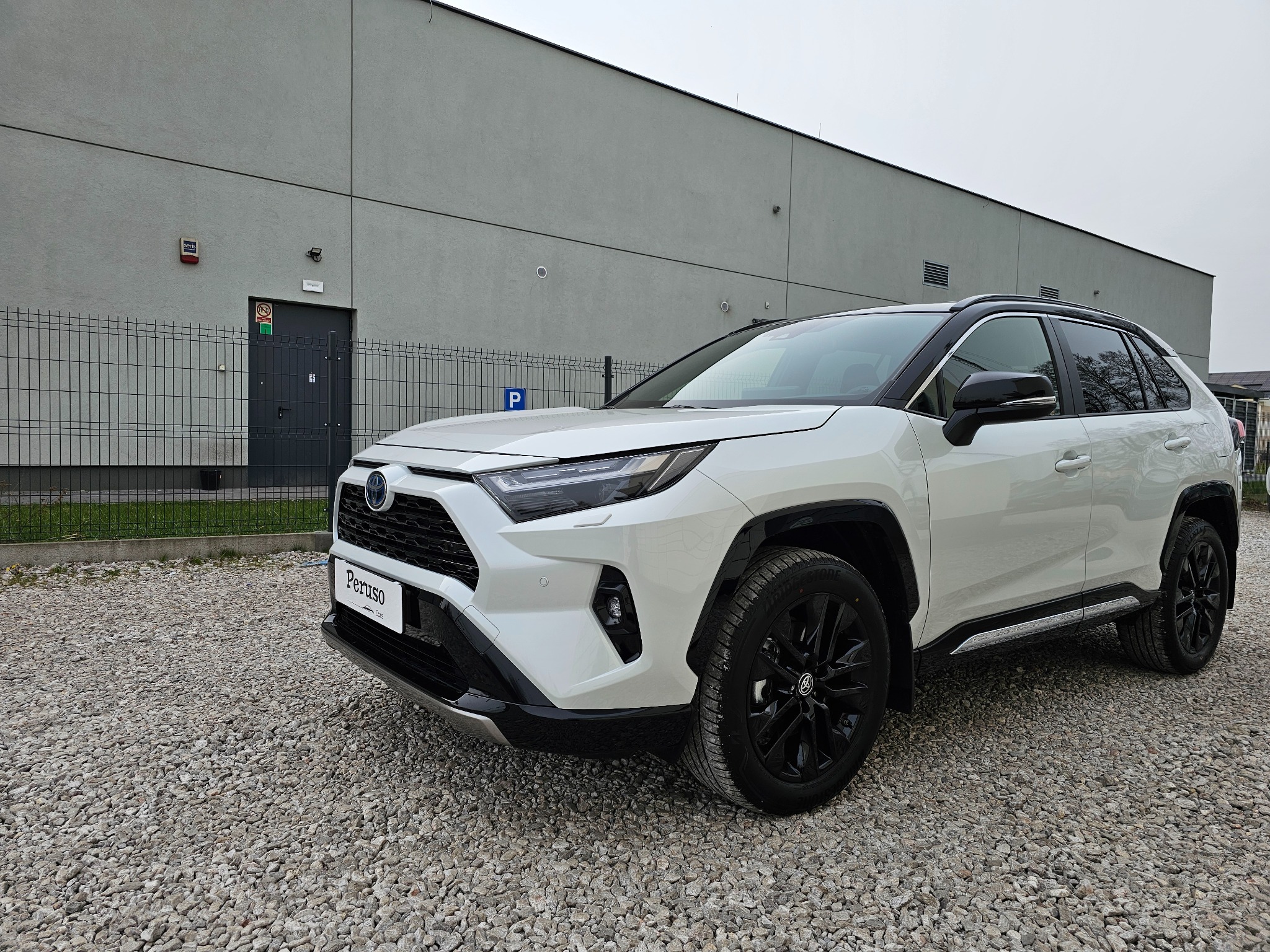 Biały samochód typu SUV, Toyota RAV4, zaparkowany na żwirowym parkingu przed szarym budynkiem magazynowym z widocznym znakiem parkingu.