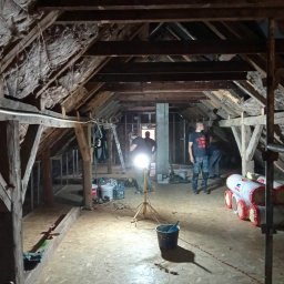 G.M Complex -Profi - Adaptacja poddasza: ekipa montuje izolację w drewnianej konstrukcji dachu. Widoczne belki, wełna mineralna, narzędzia i ekipa w trakcie pracy. Oświetlenie robocze.