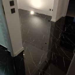 G.M Complex -Profi - Błyszcząca, czarna podłoga z białym żyłkowaniem, ułożona w kwadraty. Widoczne fragmenty ścian i drzwi. W głębi pomieszczenia kabel i lampa robocza.