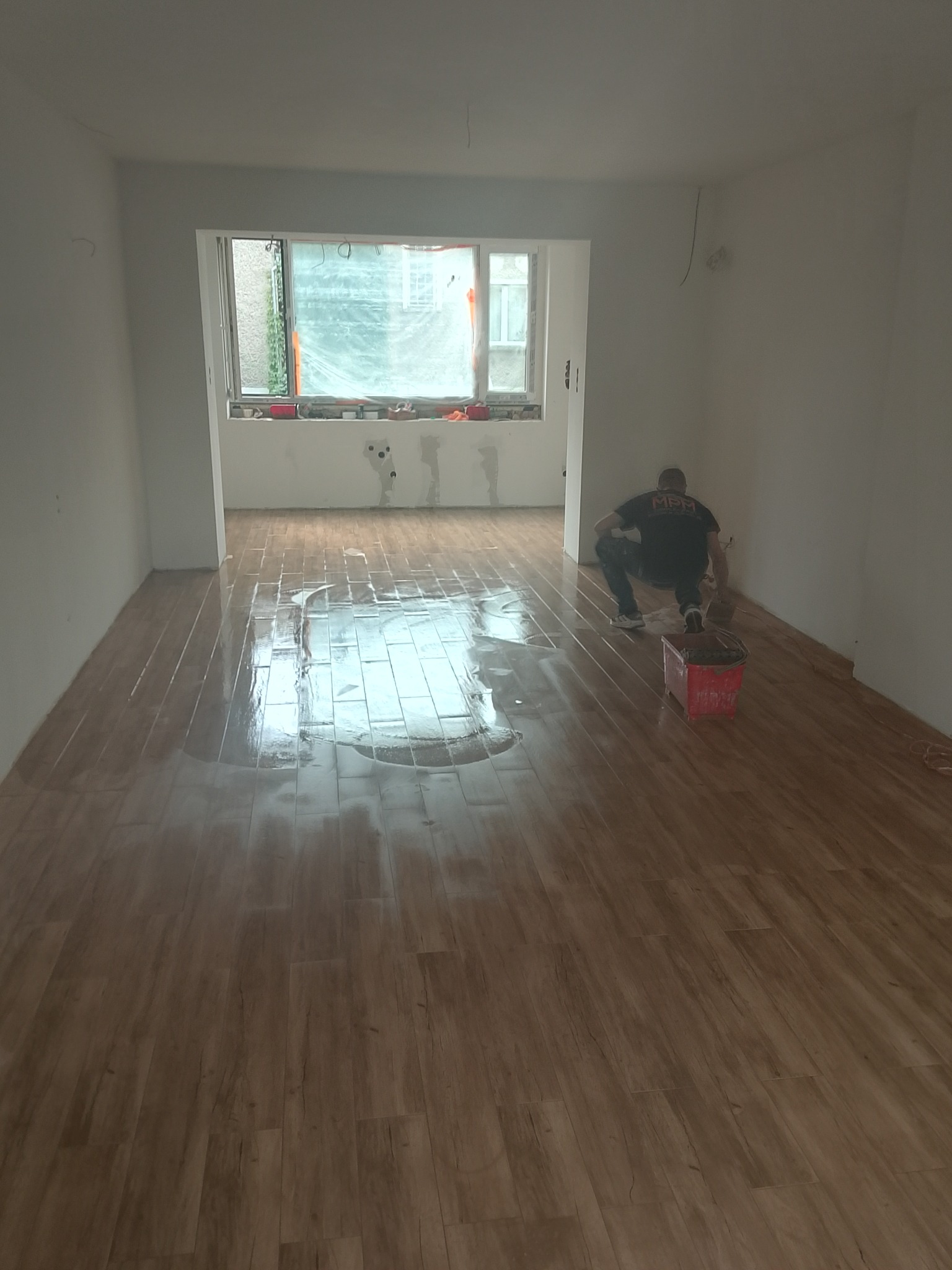 Układanie podłogi z paneli drewnopodobnych w jasnym wnętrzu. Widoczny pracownik podczas montażu. Okno przesłonięte folią ochronną.