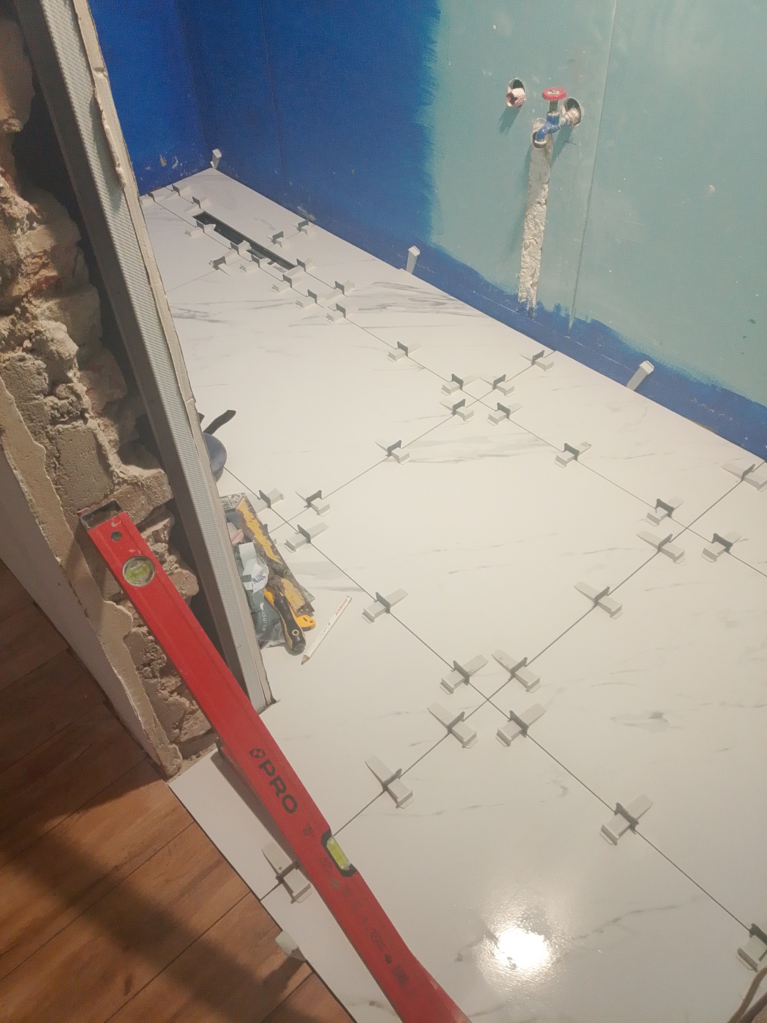 Białe płytki podłogowe z krzyżykami dystansowymi w trakcie układania. Czerwona poziomica oparta o ścianę. Częściowo wykończona ściana w tle.