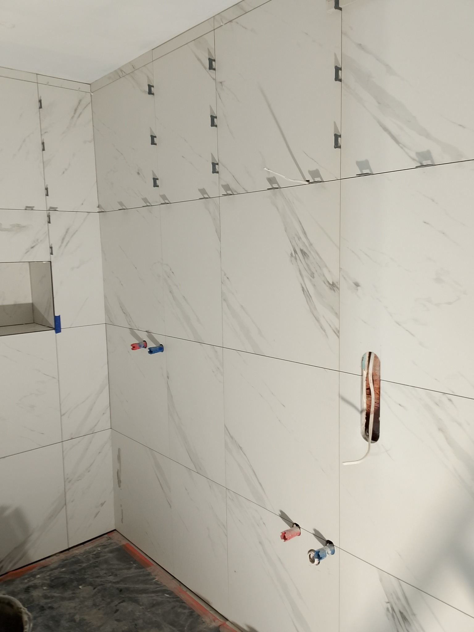 Ściana w łazience wyłożona białymi płytkami imitującymi marmur, z widocznymi krzyżykami dystansowymi, instalacją wodną i elektryczną w trakcie montażu.