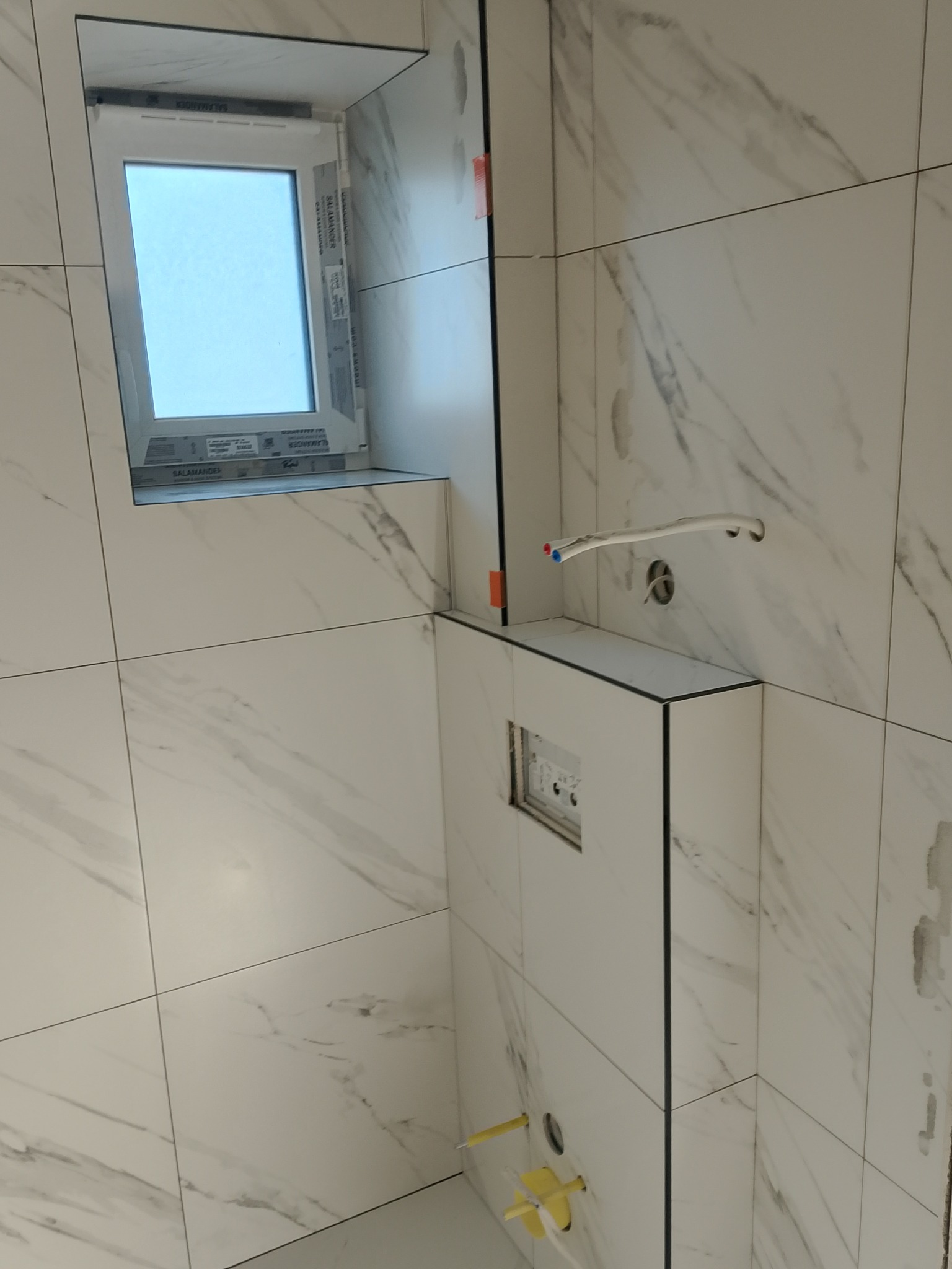 Wnętrze łazienki w trakcie wykańczania: białe płytki imitujące marmur, okno z PCV, zabudowa WC. Widoczne elementy instalacji i akcesoria montażowe.