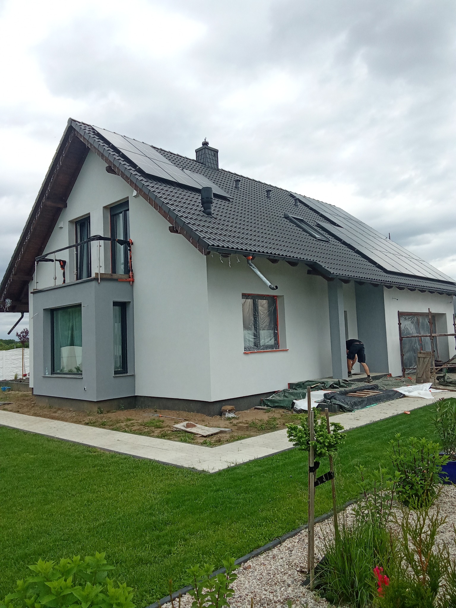 Nowoczesny dom jednorodzinny z panelami słonecznymi na dachu, z szarym wykończeniem elewacji i pracami wykończeniowymi w toku, widok z ogrodu.