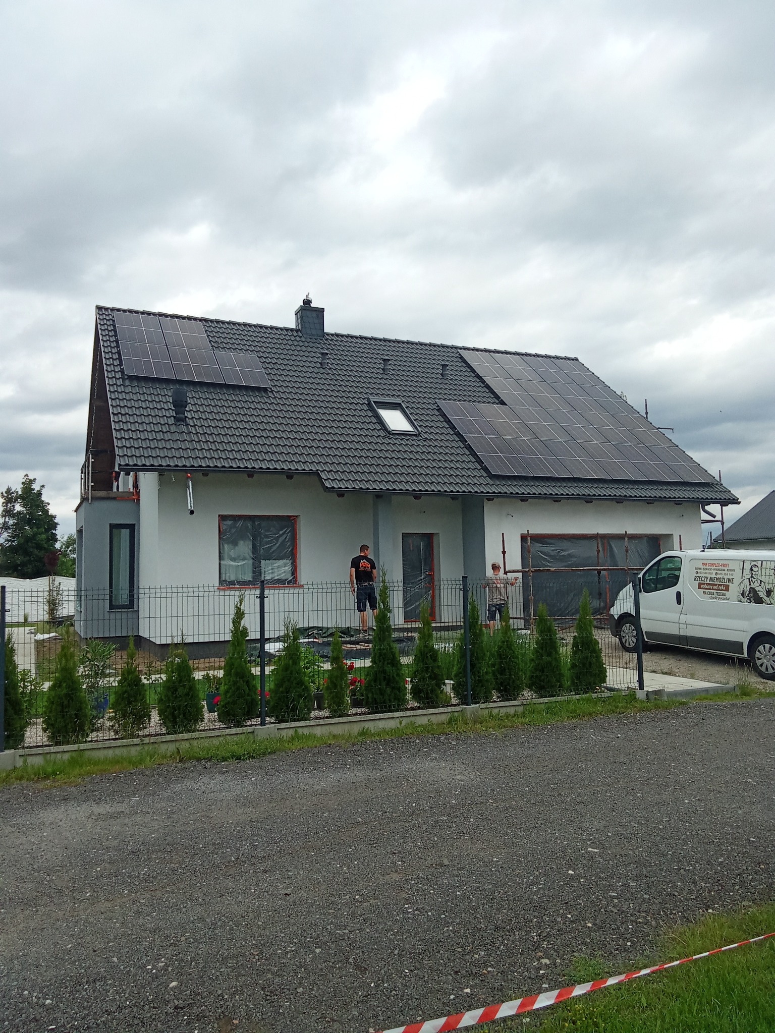Nowoczesny dom z panelami słonecznymi na dachu, widoczny zza ogrodzenia, z ekipą montującą i zaparkowanym busem firmy. Pochmurne niebo w tle.
