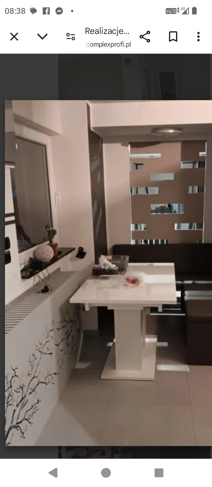 Jasne wnętrze z białym, rozkładanym stołem i dekoracyjną ścianą z motywem drzewa. W tle widoczna ściana z geometrycznymi wycięciami i ciemna kanapa.