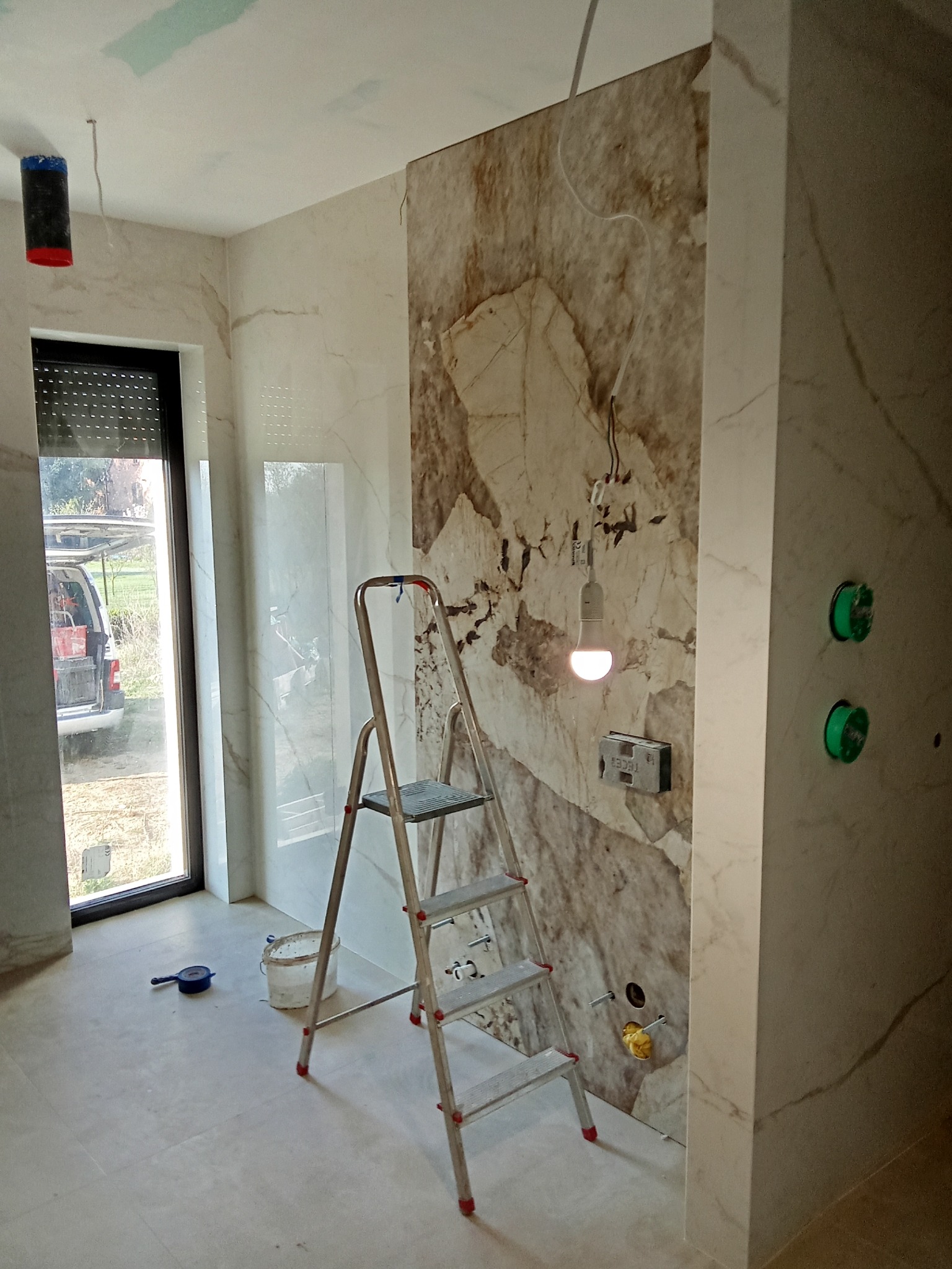 Wnętrze łazienki w trakcie wykańczania: marmurowe płytki na ścianach, drabina,  żarówka na kablu, widoczne instalacje. Detal:  okno z żaluzjami i widok na zewnątrz.