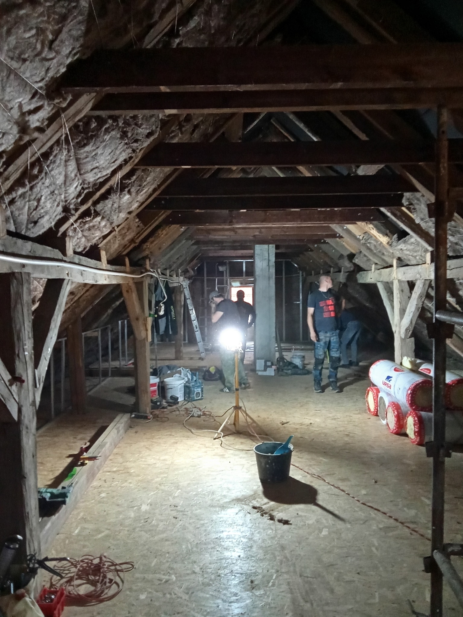 Adaptacja poddasza: ekipa montuje izolację w drewnianej konstrukcji dachu. Widoczne belki, wełna mineralna, narzędzia i ekipa w trakcie pracy. Oświetlenie robocze.