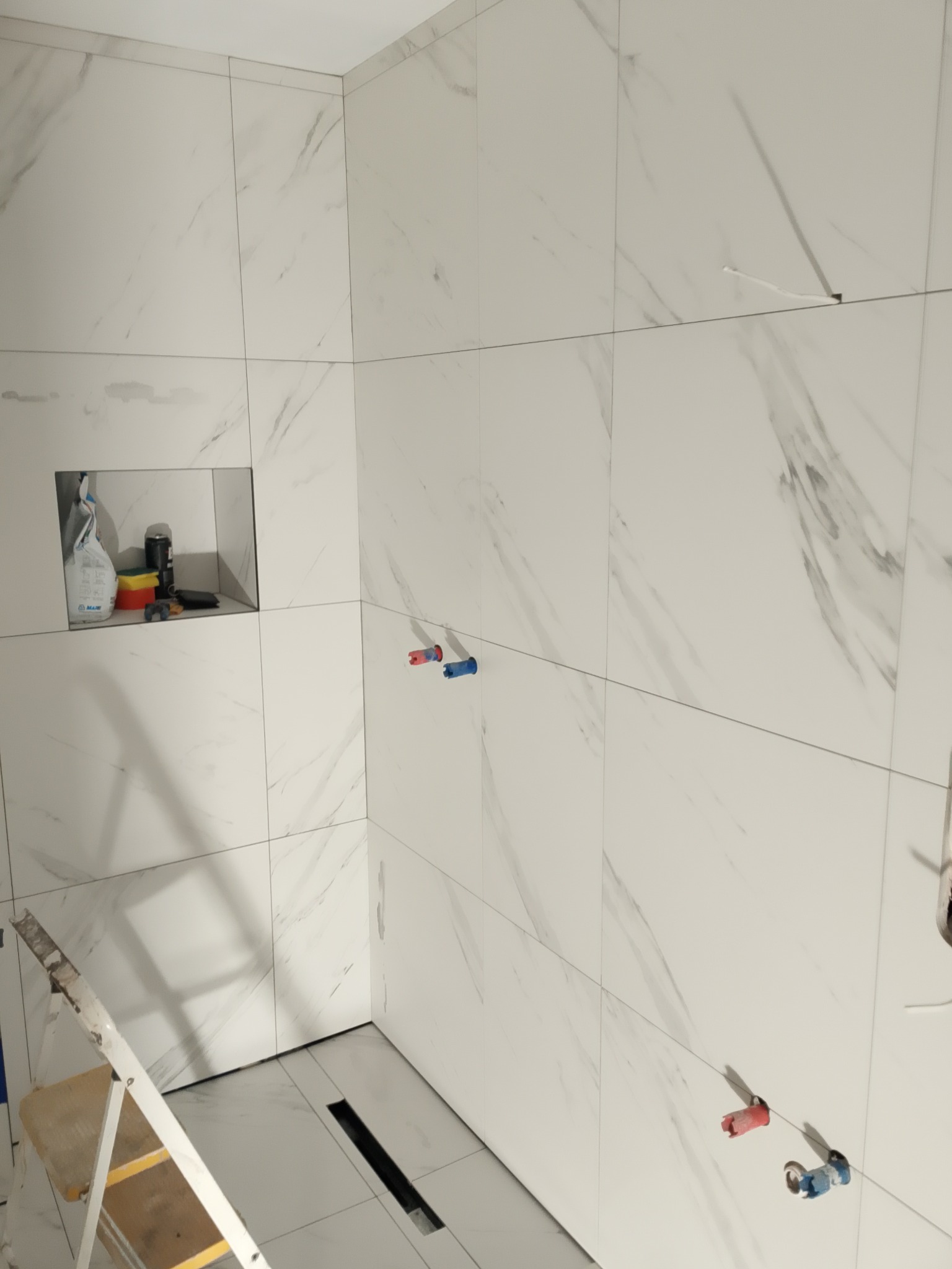 Łazienka w trakcie wykańczania: białe płytki imitujące marmur, odpływ liniowy, drabina malarska. Widoczna wnęka z materiałami budowlanymi.