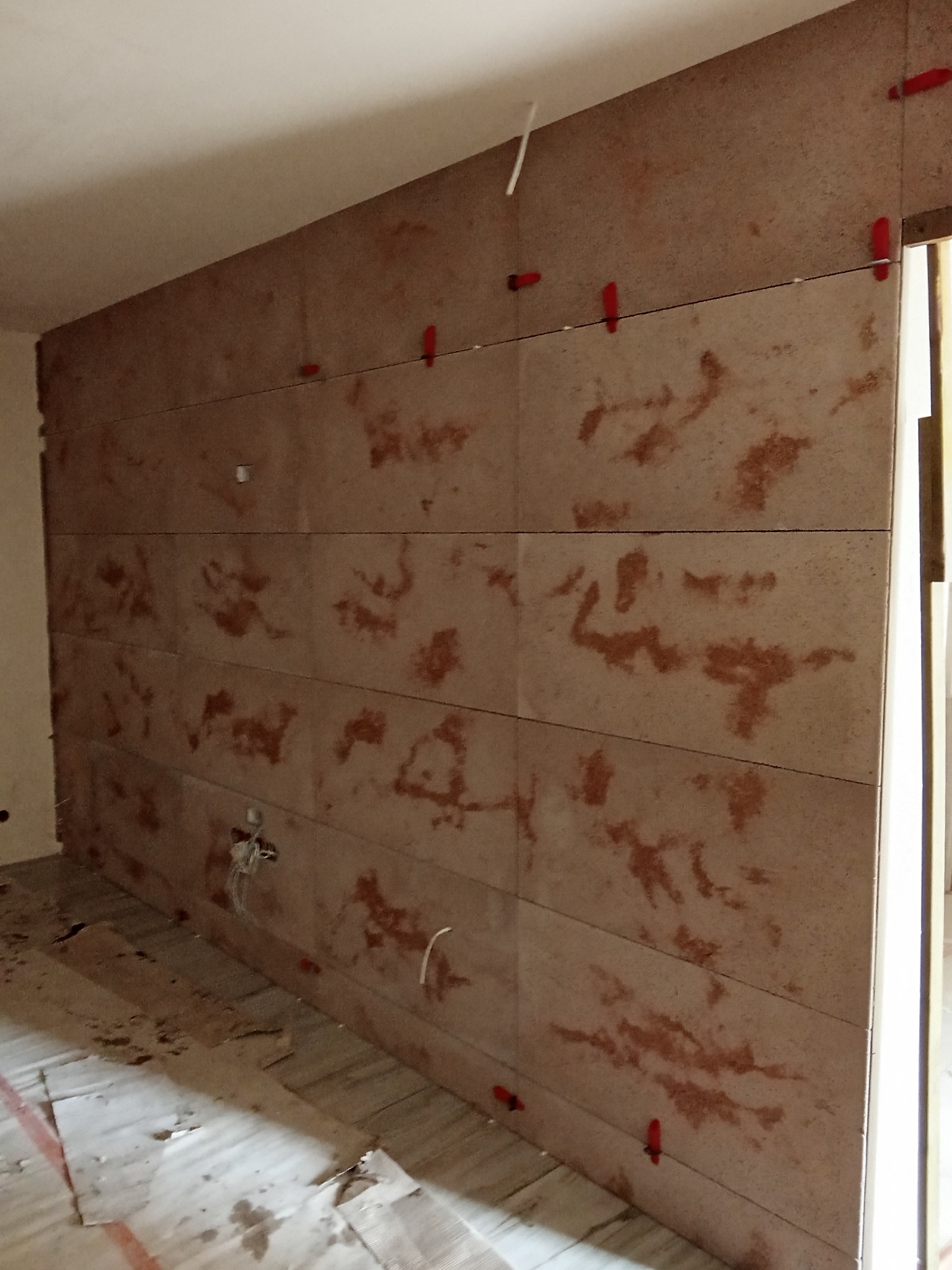 Montaż paneli ściennych z użyciem czerwonych klipsów poziomujących. Widoczne kable elektryczne wystające ze ściany. Podłoga zabezpieczona kartonem.