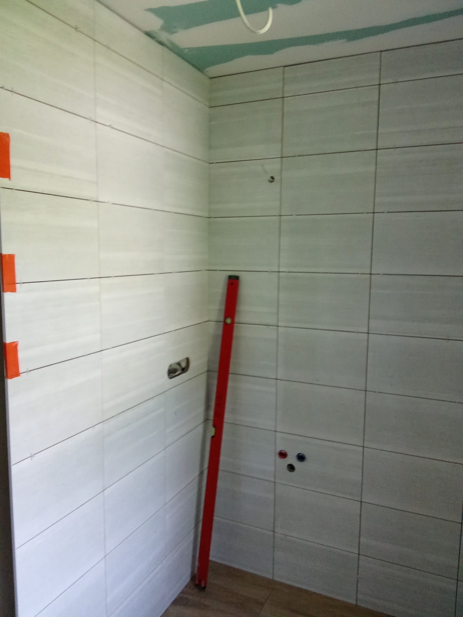 Ściana łazienki z ułożonymi, poziomymi, białymi płytkami. Czerwony poziomica oparta o ścianę. Widoczne otwory na instalacje. Na górze surowy sufit.