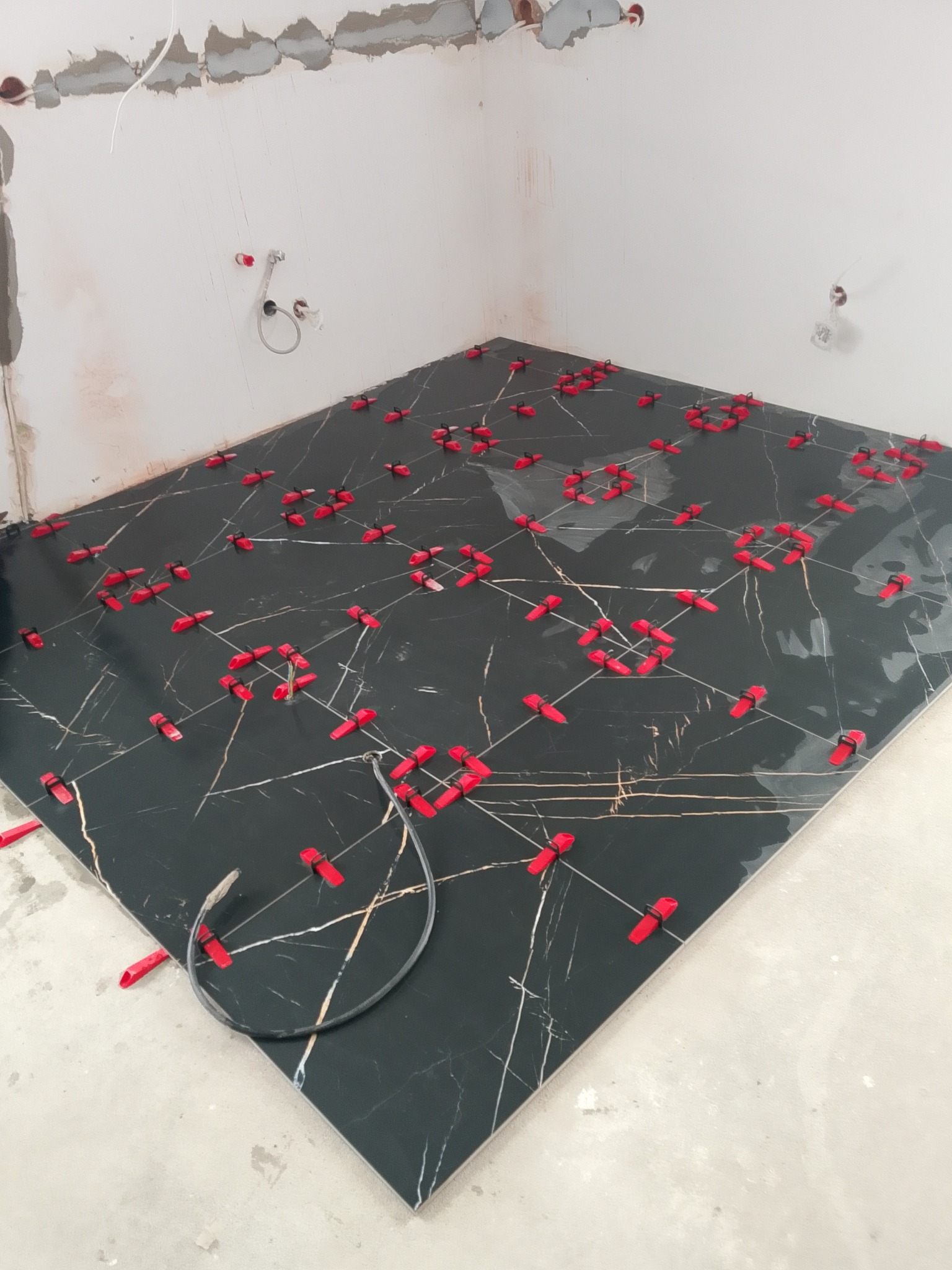 Układanie czarnych płytek podłogowych z białym wzorem żyłkowania, z użyciem czerwonych klipsów poziomujących. Widoczne surowe ściany w trakcie remontu.