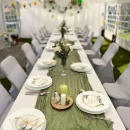 Udekorowany stół w białym namiocie: białe krzesła, zielony bieżnik, kwiaty w wazonie, świece i talerze z dekoracjami. Balony i dekoracje na suficie.