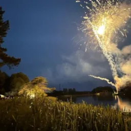Nocne niebo rozświetlone fajerwerkami nad jeziorem, w tle widoczny namiot i sylwetki ludzi, tworząc atmosferę imprezy plenerowej.