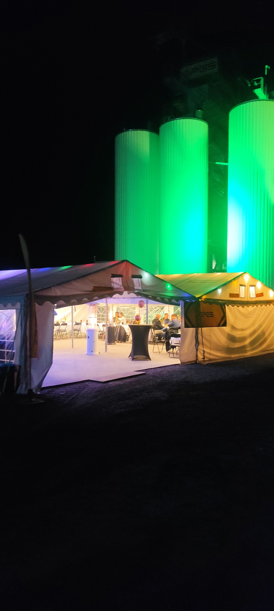 Dwa oświetlone namioty eventowe z gośćmi w środku, na tle trzech jasnozielonych, podświetlonych silosów. Ujęcie nocne, z niskiej perspektywy.