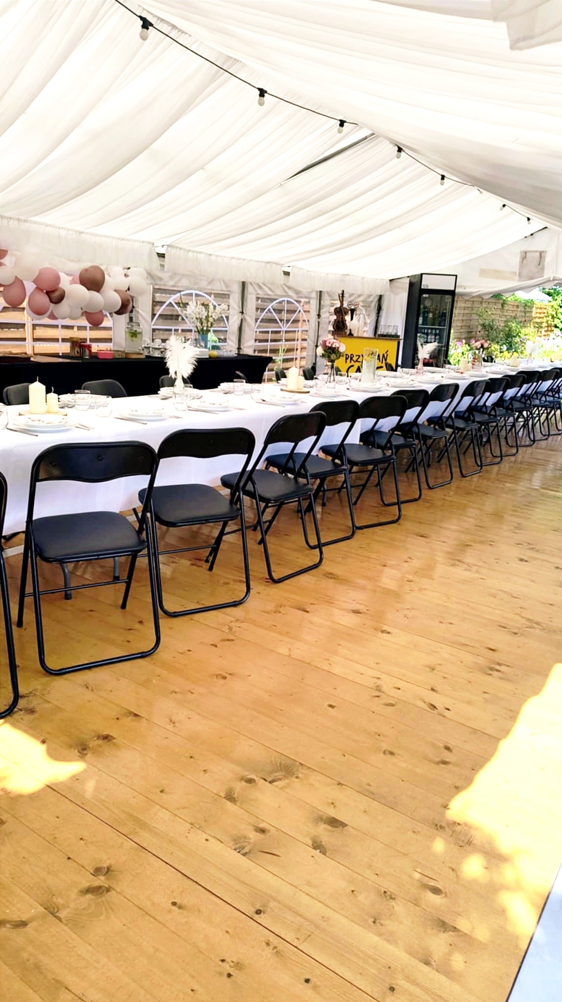 Uroczysty biały namiot z rzędem czarnych krzeseł, stołem z zastawą i dekoracjami. Drewniana podłoga, balonowa girlanda i bufet w tle.