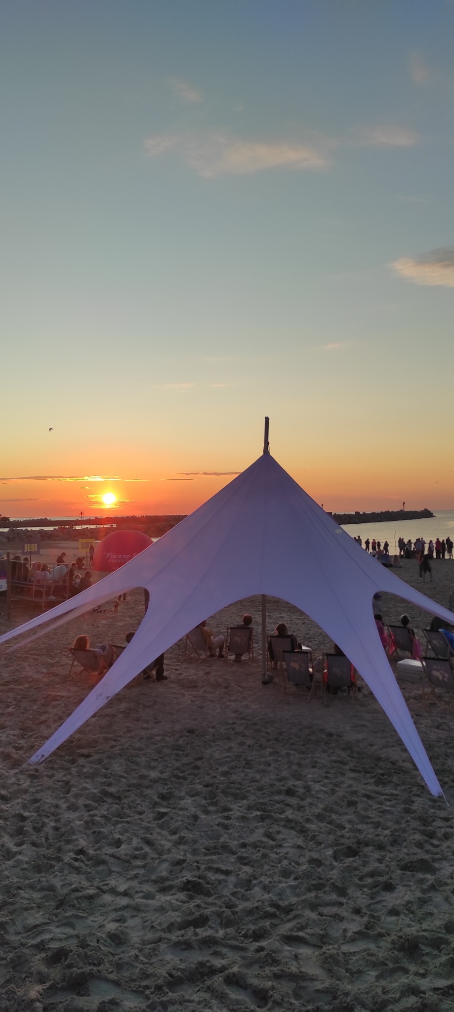 Plażowy namiot w kształcie gwiazdy na tle zachodzącego słońca. Widoczni ludzie siedzący w leżakach. Romantyczna atmosfera, idealna na event na plaży.