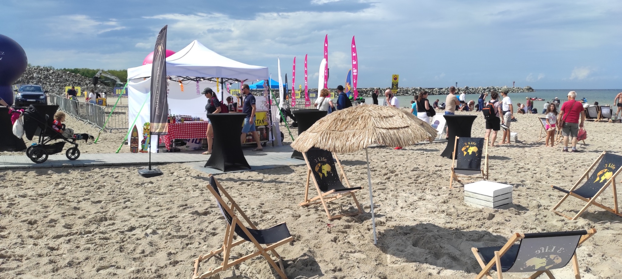 Plaża z leżakami, parasolem z trzciny i stoiskami eventowymi w tle. Widoczni ludzie i nadmorski krajobraz.