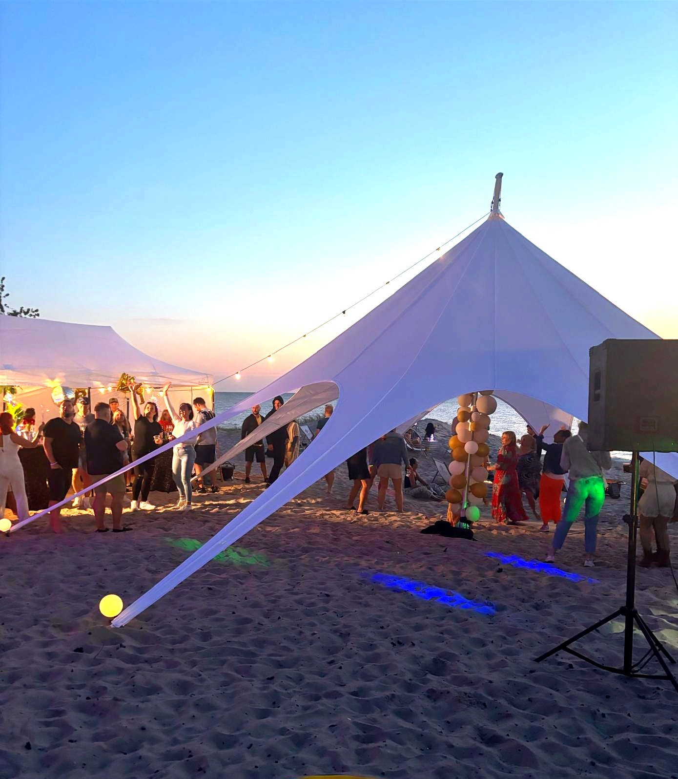 Plażowa impreza pod białym namiotem w kształcie gwiazdy, oświetlonym kolorowymi światłami. W tle zachód słońca i bawiący się ludzie.