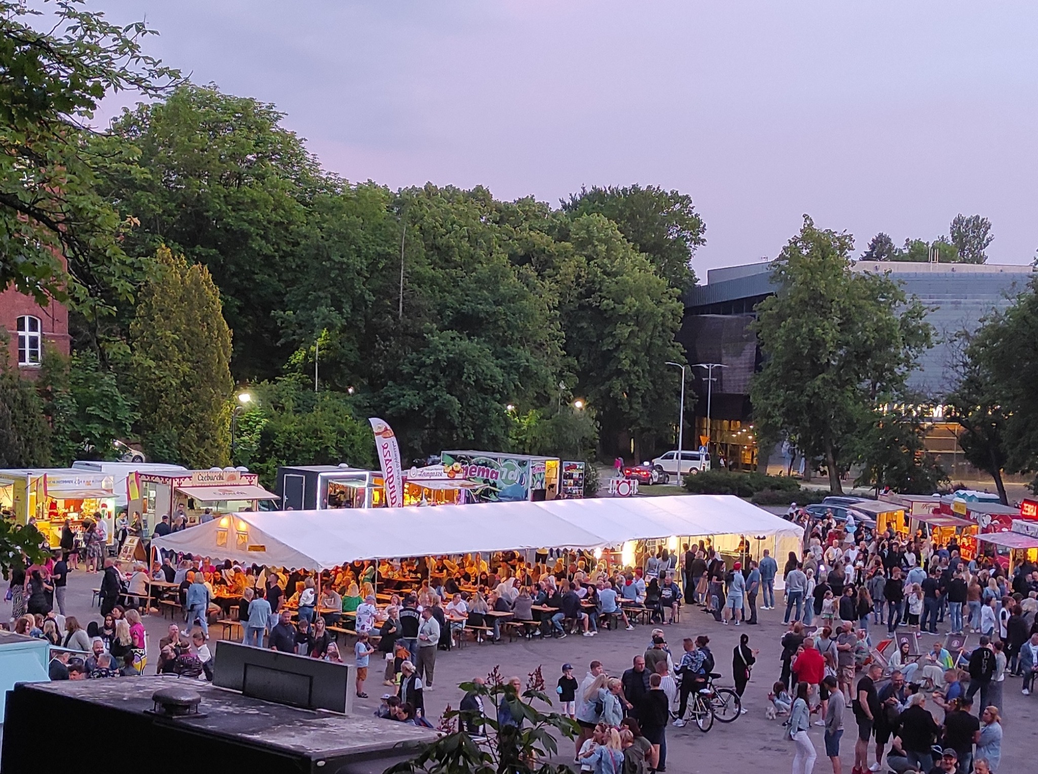 Zadaszony biały namiot z oświetleniem, tłum ludzi siedzących przy drewnianych stołach, food trucki w tle, wieczorna atmosfera, drzewa i budynki w tle.