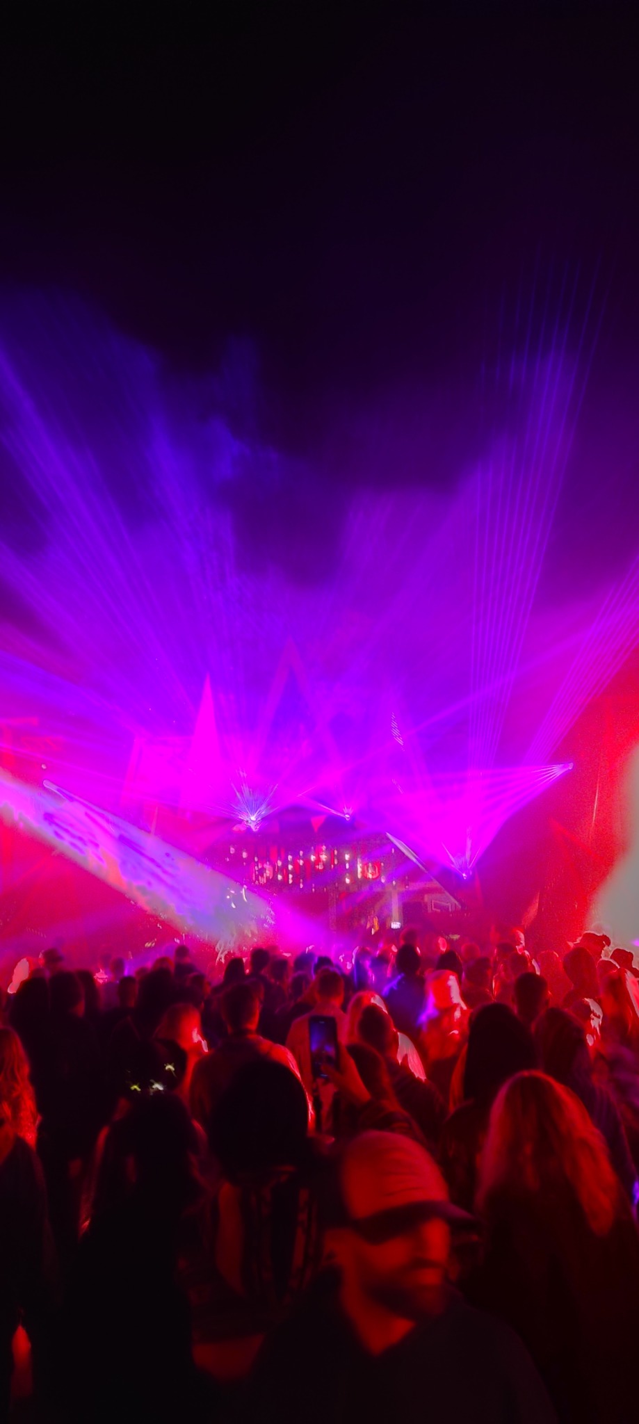 Tłum ludzi na koncercie oświetlonym intensywnymi, purpurowo-czerwonymi laserami. W tle scena z elementami dekoracyjnymi. Ujęcie z dołu, dynamiczne.