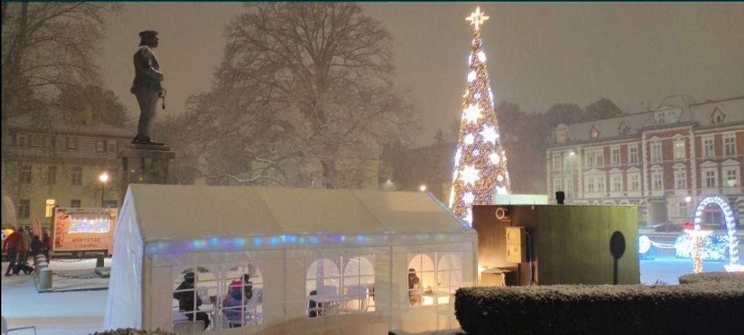 Zimowy pejzaż miejski: biały namiot z oświetleniem na tle ośnieżonego rynku z pomnikiem, choinką i budynkami. W namiocie widoczni ludzie.