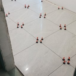 Marcin Polek Usługi Remontowo-Serwisowe MM - Jasnoszare płytki podłogowe ułożone z użyciem systemu poziomowania płytek (klipsy i kliny), widoczne surowe ściany i okno.