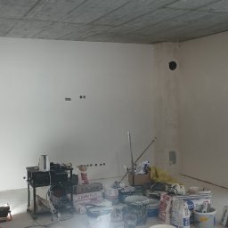 Marcin Polek Usługi Remontowo-Serwisowe MM - Wnętrze w stanie deweloperskim w Bochni, ściany po nałożeniu gładzi, widoczne worki z zaprawą, narzędzia i prowizoryczne oświetlenie podłogowe.