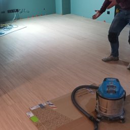 Marcin Polek Usługi Remontowo-Serwisowe MM - Układanie paneli podłogowych w pomieszczeniu z pomalowanymi na zielono ścianami, widoczny mężczyzna wskazujący na podłogę, odkurzacz przemysłowy i kostka styropianu.