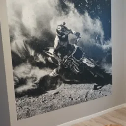 Czarno-biała fototapeta na ścianie przedstawiająca motocyklistę w kasku podczas jazdy w terenie, widoczny fragment krzesła i stołka na tle paneli podłogowych