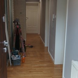 Marcin Polek Usługi Remontowo-Serwisowe MM - Korytarz w trakcie prac wykończeniowych, widoczna drabina malarska z narzędziami, ściany pomalowane na szaro, drewniana podłoga przechodzi w białe płytki, oświetlenie sufitowe punktowe.